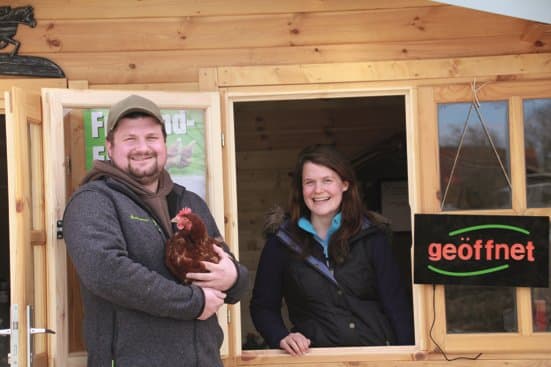 Anne Wember-Mersmann und ihr Mann vor dem geöffneten Verkaufshaus, er hält ein Huhn im Arm